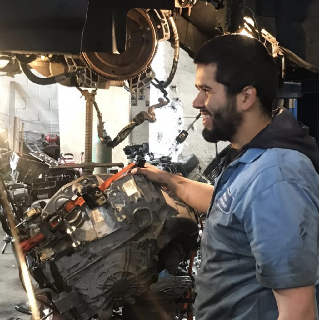 A Bob's Auto Repair Mechanic Working On A Hanging Transmission And Smiling